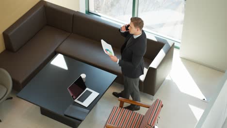 businessman in office talking on phone holding a document with colorful charts analysis, man in suit having business smartphone communication in modern meeting room - high angle wide view