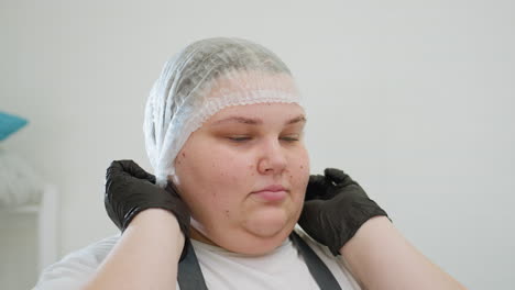 beautician wearing black gloves carefully puts on disposable shower cap in preparation for a beauty procedure. captured in clean, white setting emphasizing hygiene and professional readiness