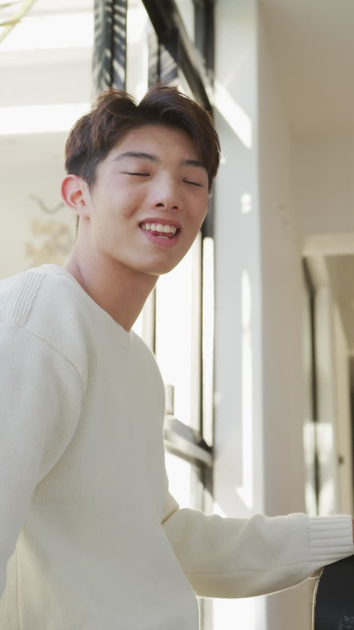 거실에서 스케이트보드와 함께 미소 짓는 아시아 남성 십대 초상화의 수직 비디오