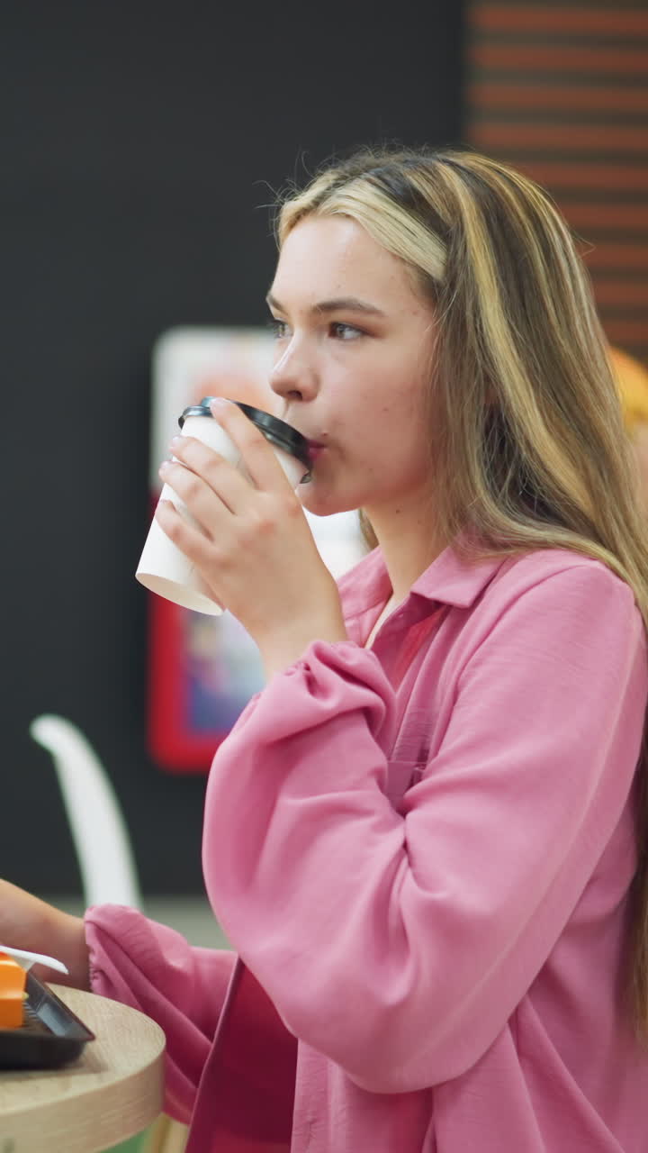 dama de vestido rosa recoge su bebida de la mesa, lo huele, y toma un sorbo con una expresión satisfecha, papas fritas y una hamburguesa se colocan en una bandeja en la mesa, otras personas están sentadas alrededor