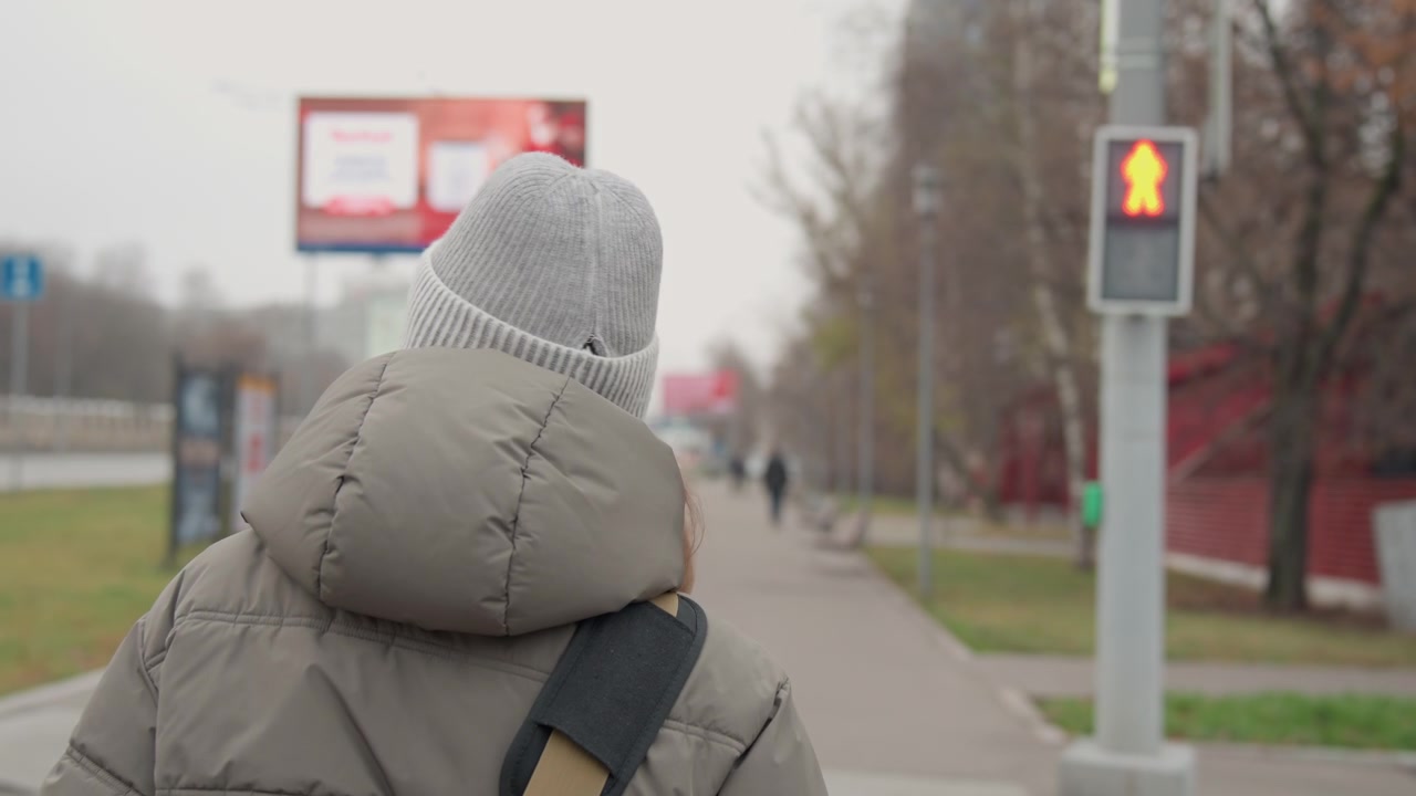 Mulher branca caminhando na calçada perto da faixa de pedestres, passageira parada no sinal vermelho de pedestres em uma manhã urbana nublada, jaqueta puffer, gorro de tricô e bolsa de ombro transmitem rotina tranquila e reflexiva.