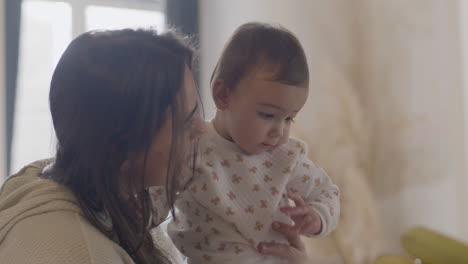 happy woman holding her baby daughter and talking to her while sitting at kitchen table on the morning