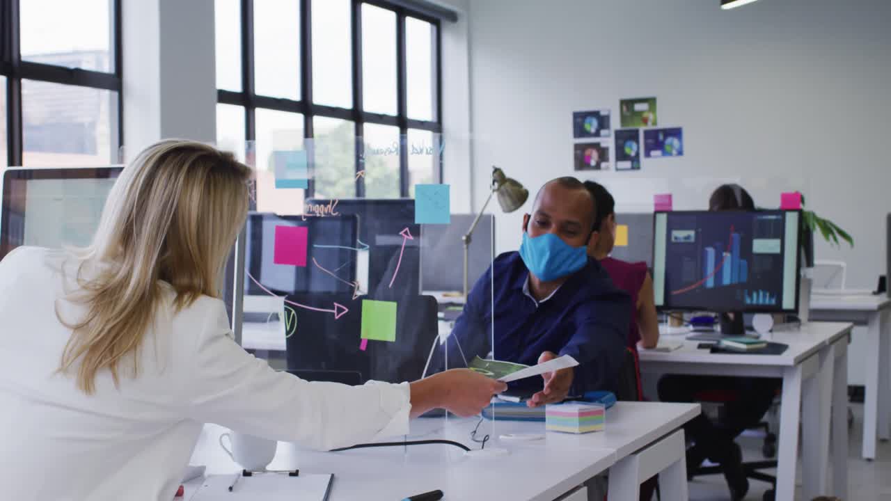 Diversos compañeros de trabajo con mascarillas usando computadoras y pasando documentos en la oficina.