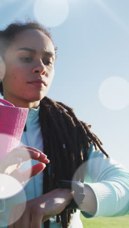 Multiple-white-spots-floating-against-african-american-fit-woman-using-smartwatch-at-sports-field