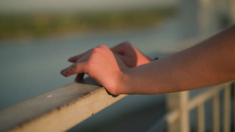 close-up of white person's hand resting on iron railing during sunset, fingers lightly touching, with blurred background featuring greenery in the distance