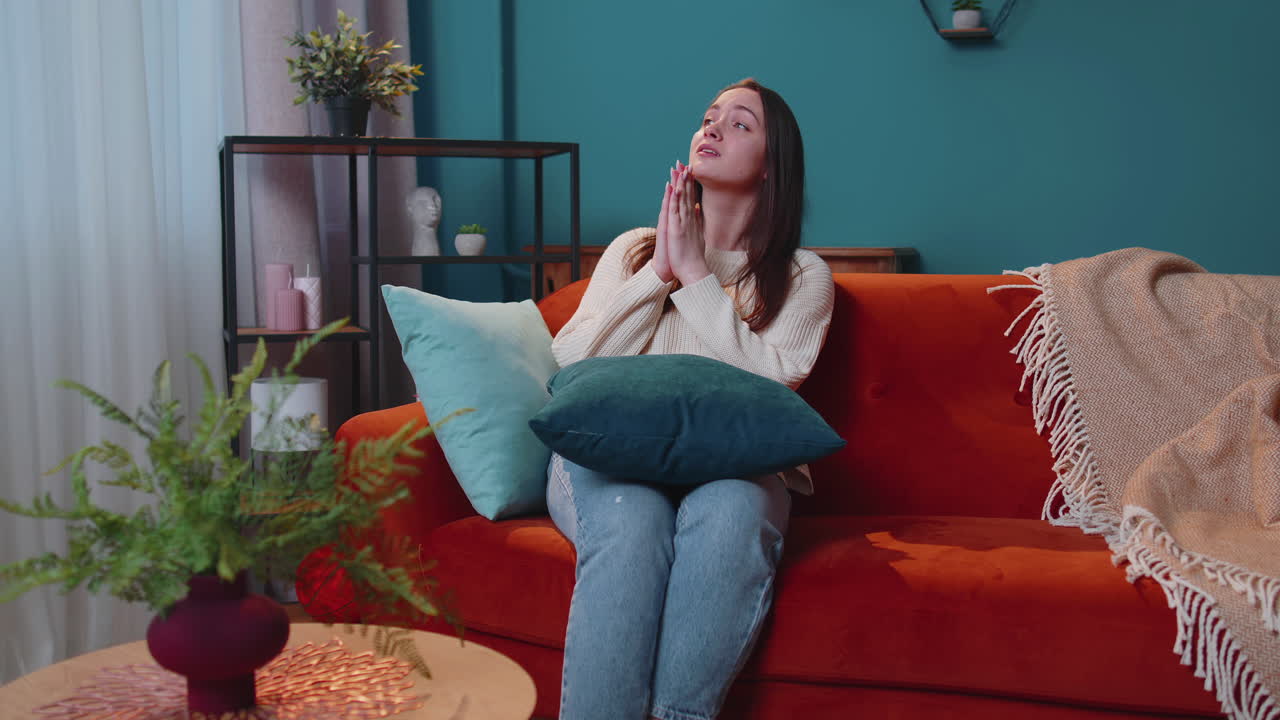 슬픈 젊은 성인 여성은 집에서 소파에 앉아 끔찍한 소식에 충격을 받고 기도하고 도움을 요청합니다.