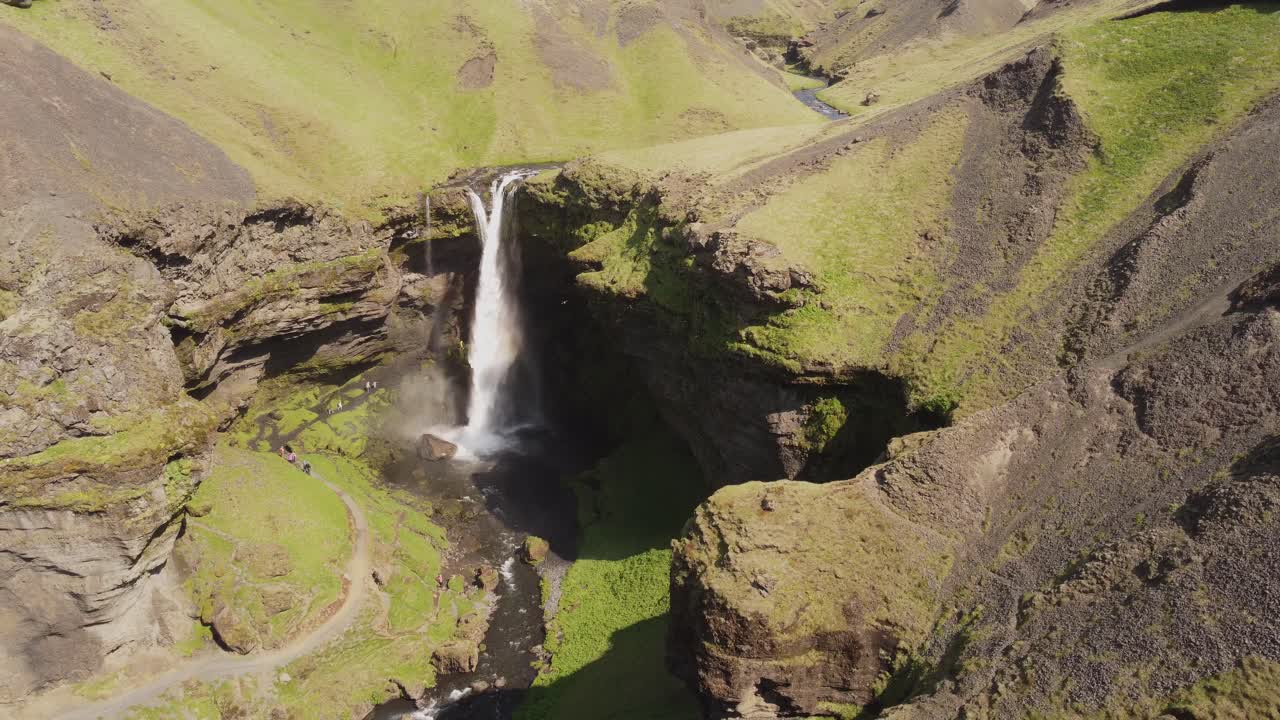 toma aérea de gran angular de una hermosa cascada en islandia en un día soleado con pájaros volando y un pequeño arco iris frente a los acantilados y rocas verdes y cubiertos de musgo