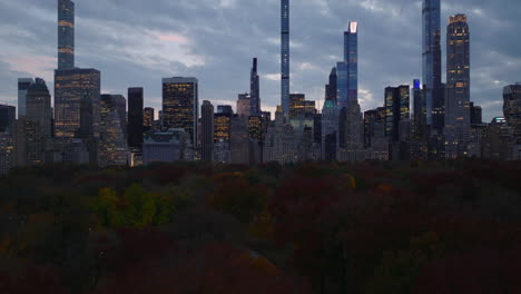 Slide-and-pan-footage-of-downtown-skyscrapers-and-high-rise-office-buildings-against-twilight-sky.-Low-flight-above-autumn-Central-park.-Manhattan,-New-York-City,-USA