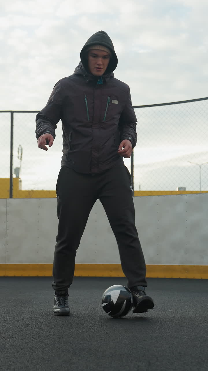 Man in hoodie practicing soccer drills on sport arena, demonstrating footwork and ball control under daylight, background shows lamp pole, urban residential buildings, and goalpost on a fenced court