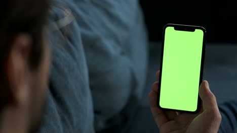 a young man is lying on a sofa in a room at home, looking at the camera shows a smartphone with a green screen integration of a new application