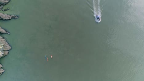 vista aérea de arriba hacia abajo sobre tres kayakistas en aguas tranquilas y turbias mientras un barco pesquero motorizado pasa a su izquierda.