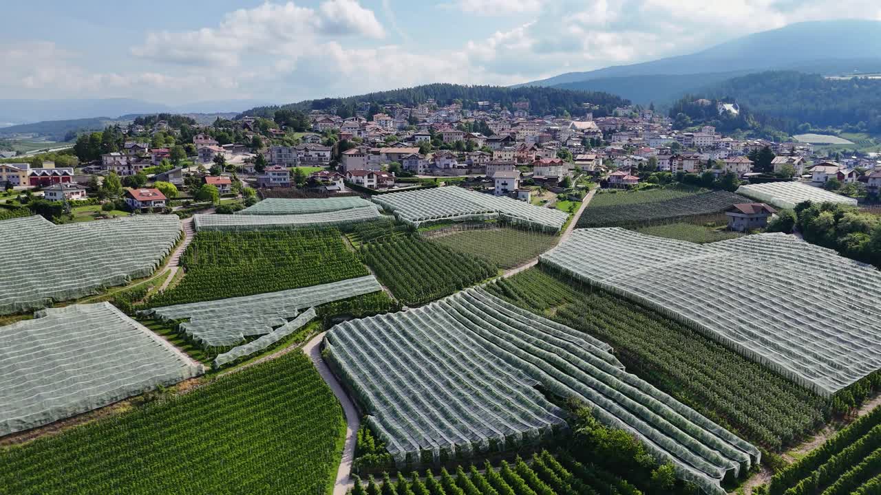 Drone footage captures Coredo’s greenhouses and farmland in a slow circle motion, with a village and hills under a partly cloudy sky