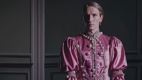 un joven noble con un traje de satén rosa históricamente auténtico y collares de perlas de pie con confianza en un interior adornado de paneles gris oscuro, que representa la elegancia aristocrática del renacimiento