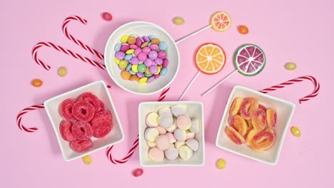 white bowls filling with sweet gummy and chocolate candies and lollypops on pastel pink background. stop motion animation flat lay