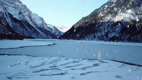 Winter-mountains-and-frozen-lake-surface-during-snowfall,-aerial-fly-backward-view