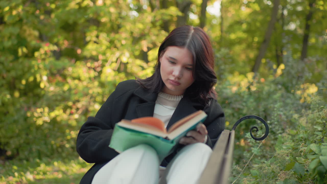 공원 벤치에 다리를 올리고 앉아 초록색 책을 읽고 있는 외로운 소녀, 차분한 표정, 페이지를 넘기면서 머리를 정리하고, 땅에는 종이컵이 놓여 있고, 가을 햇살 아래 무성한 잎이 있는 배경, 조용한 야외 학습 순간