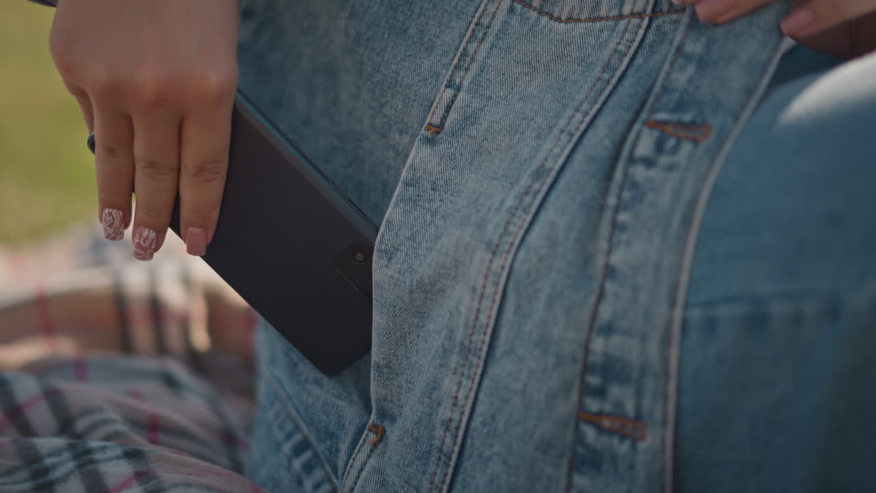 Primo piano di una mano con giacca di jeans che riposa su una coperta a quadri, unghie dipinte, telefono che viene infilato in tasca durante un picnic all'aperto, calda luce del sole, stile di vita casual, atmosfera estiva accogliente, gesto rilassato.