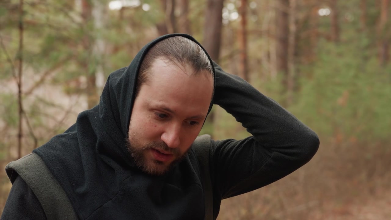 Homme barbu portant un pull en tricot à capuche sombre fait une pause dans une forêt de pins, lève la main à l'arrière de sa tête, puis adopte une posture pensive tout en regardant vers l'avant à travers une douce lumière naturelle.