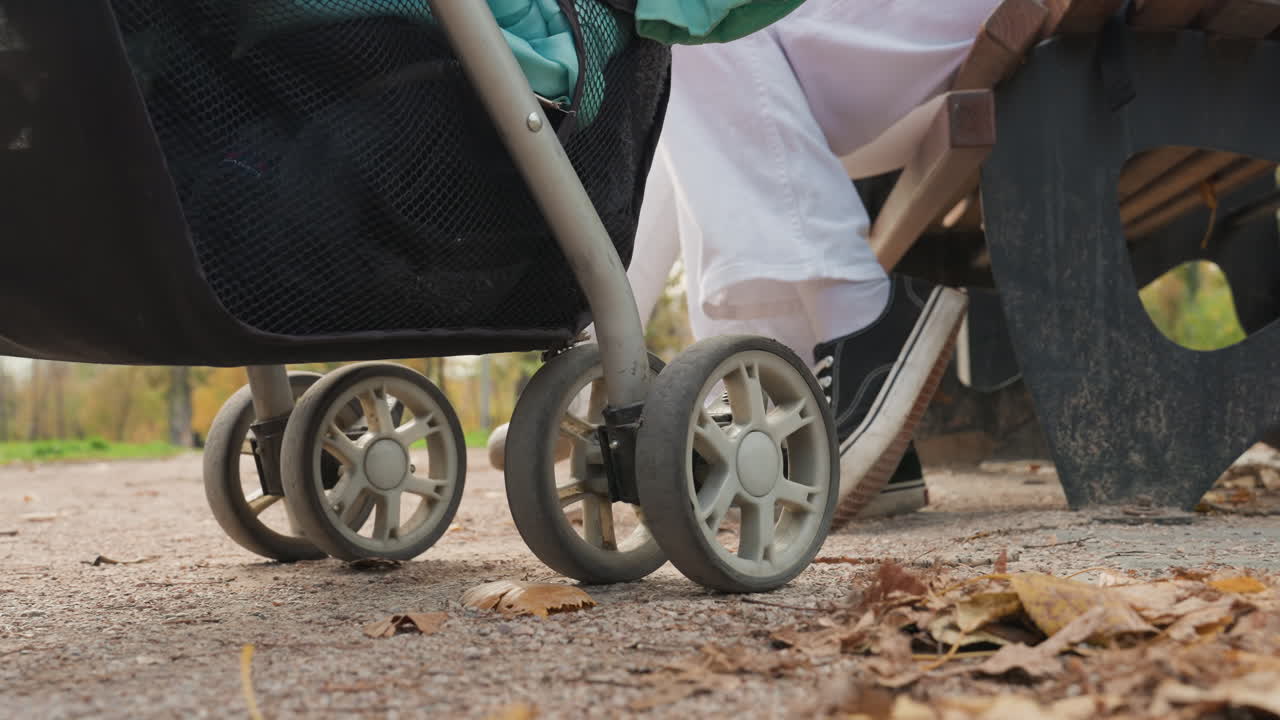 Mamma skjuter barnvagn på en stig. Närbild på hjulen på en lövtäckt stig. Lugn familjepromenad fångad i mjukt höstsolsken. Intimt familjeögonblick när mamma promenerar med bebis under höstlöven.