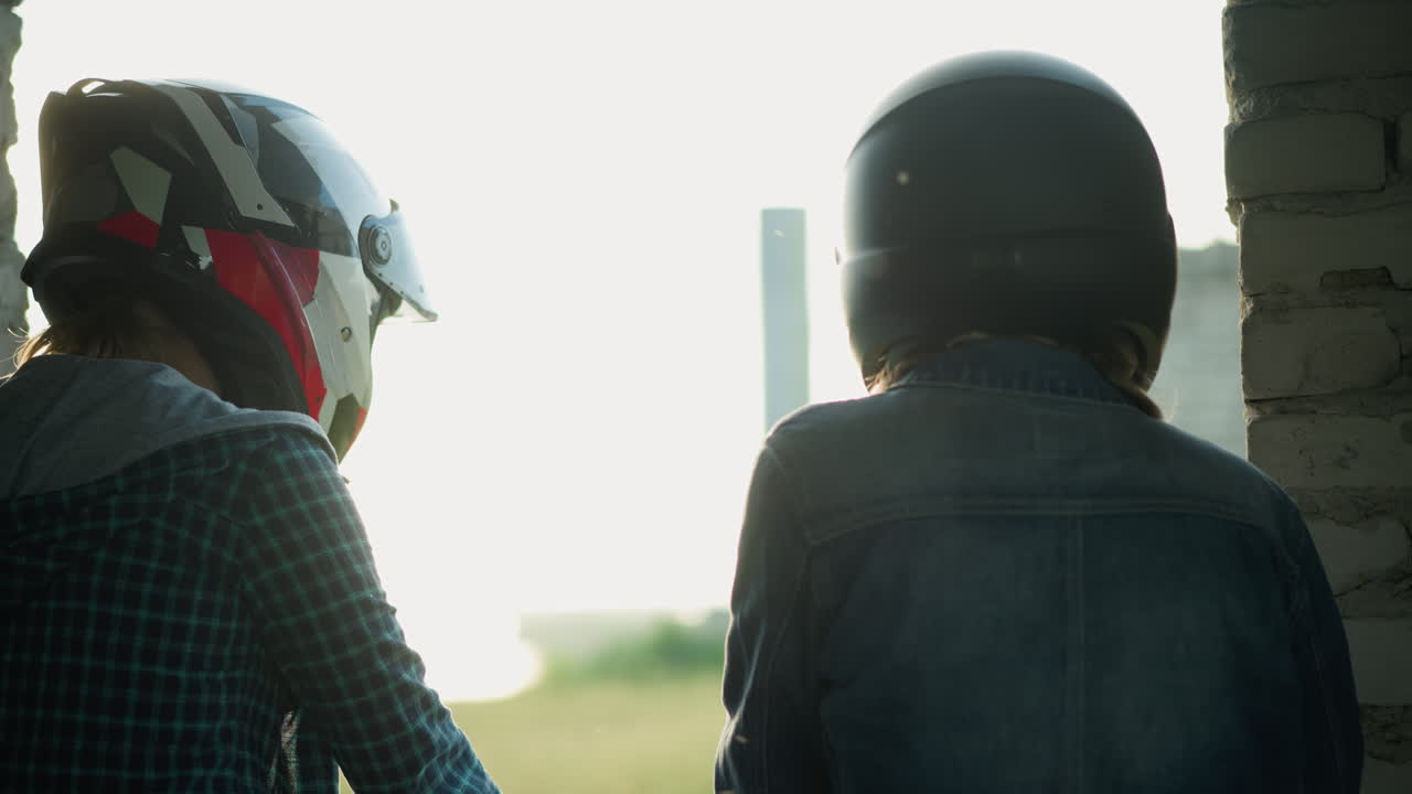 vista trasera de dos mujeres de pie cerca de una ventana, una mirando contemplativa con un fondo de edificio borroso, ambas llevan cascos, la luz crea reflejos suaves