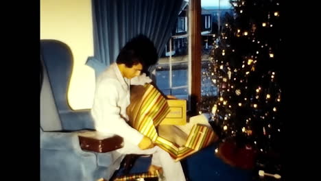 Boy Sitting on Chair Holding Golden Gift. CIRCA USA - 1970s: A boy from a 1970s video archive in the USA sitting on a chair while holding a golden gift.
