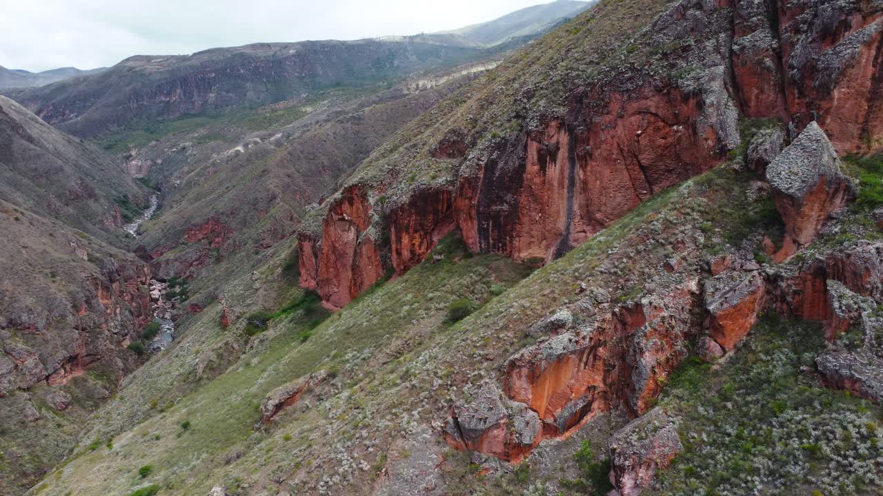 Ripresa statica con drone di un maestoso paesaggio montano caratterizzato da faraglioni rocciosi rossastri scoscesi, formazioni geologiche e un fiume tortuoso che scende attraverso una valle a canyon verde intenso sotto un…