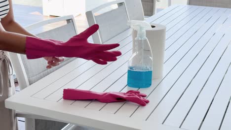person puts on pink rubber gloves at a white outdoor table with cleaning spray and paper towels, in bright natural daylight, static camera