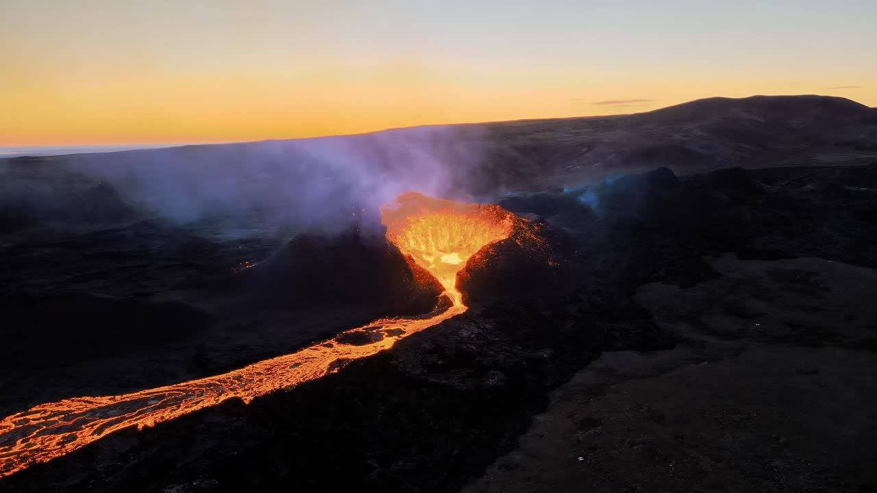 Aerial drone shot of an active volcano at sunset, showing a glowing lava river flowing across the landscape with warm evening light in the sky