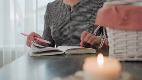 partial view of lady carefully flipping through book in hand while holding pen with candlelight burning on table basket nearby and soft natural light filtering through transparent curtain