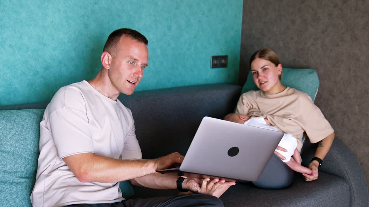 Man showing the laptop to his wife. Woman is holding a newborn and looks at laptop.
