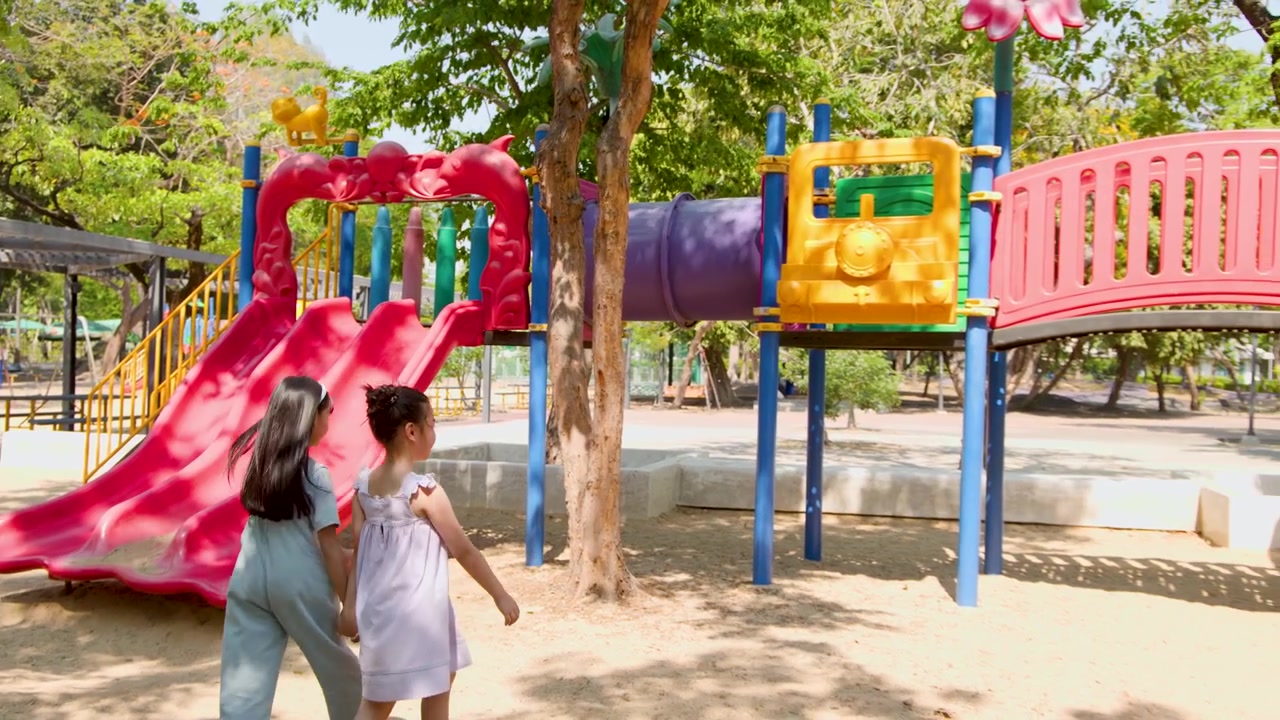 Dos niñas pequeñas se toman de la mano y pasan junto a un tobogán colorido en un parque soleado.