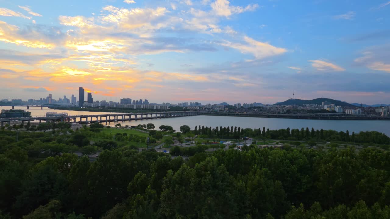An amazing aerial shot captures a color-rich sunset over the Seoul cityscape, with sunlight reflecting in the Han River, the iconic Banpo Bridge, and the lush green Hangang Park in the foreground