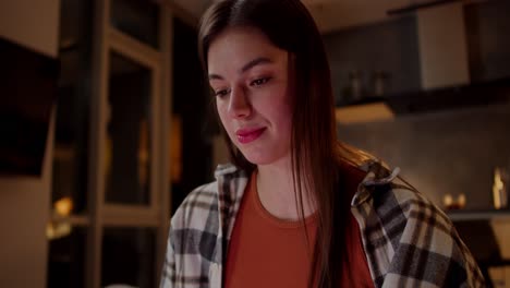 primer plano de una chica morena segura con una camisa a cuadros y una camiseta naranja viendo videos divertidos mientras está sentada en el sofá en un apartamento moderno por la noche
