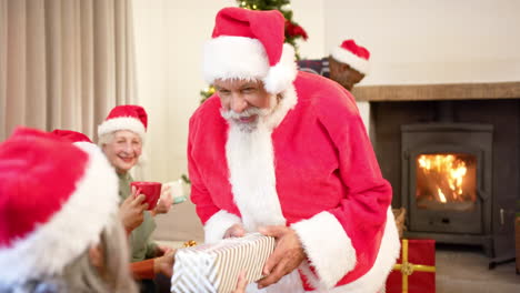 Happy-diverse-senior-friends-in-santa-outfit-and-christmas-hats-exchanging-gift-at-home,-slow-motion