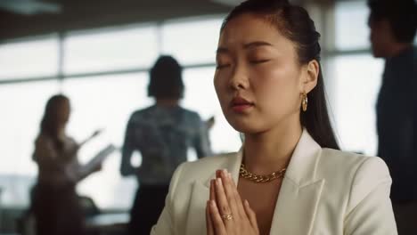 a moment of reflection: woman in office deep in thought with eyes closed, praying in the midst of a busy work environment