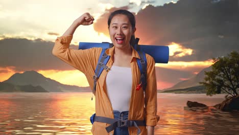 excursionista femenina asiática con mochila de montañismo sonriendo y flexionando su bíceps mientras está de pie en un lago durante el atardecer