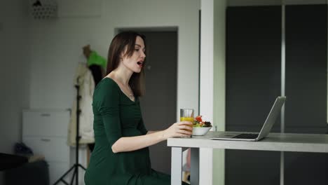 Pregnant-woman-sitting-on-the-kitchen-eats-salad-while-having-a-video-call-on-laptop
