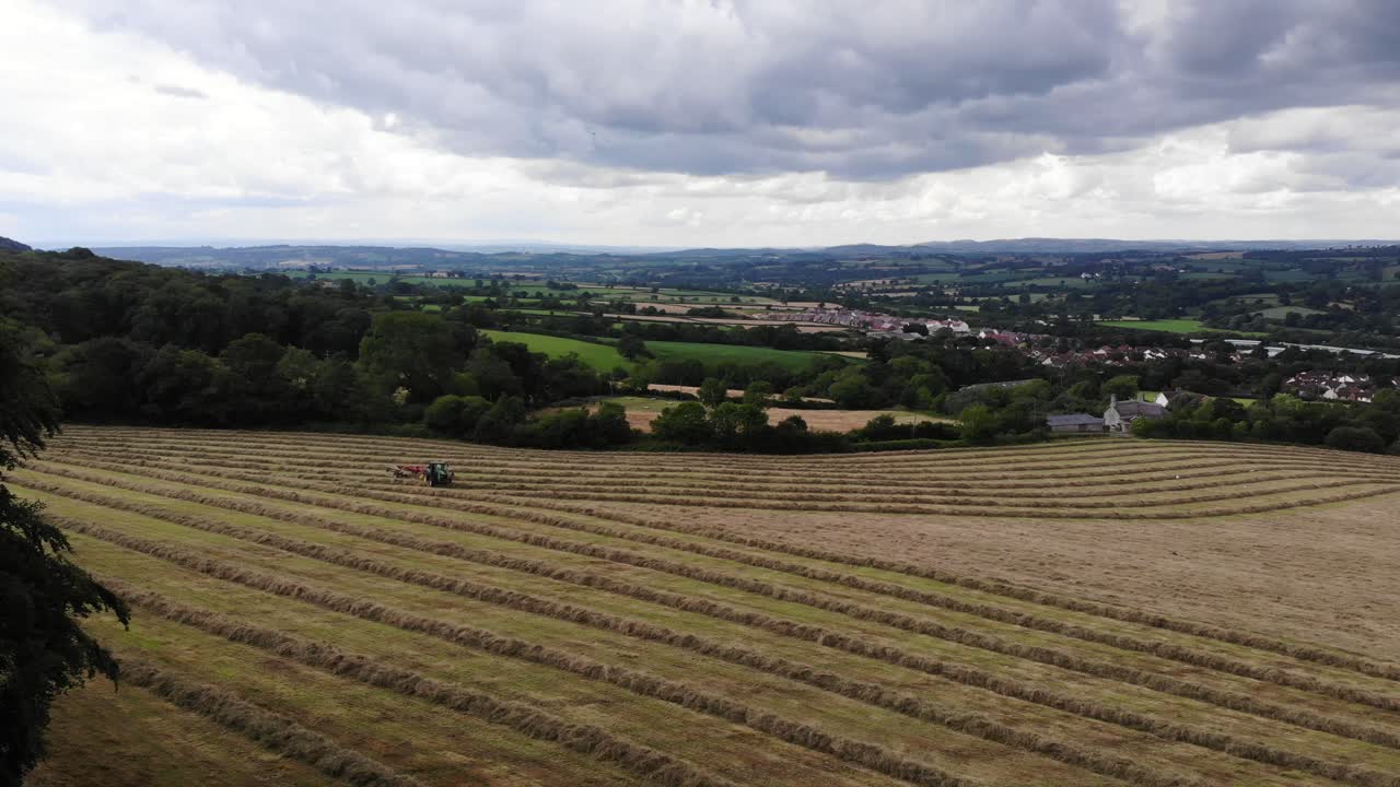 Aerial panning right shot of a tractor gathering hay on a large field with a view beyond filmed in Devon South West England