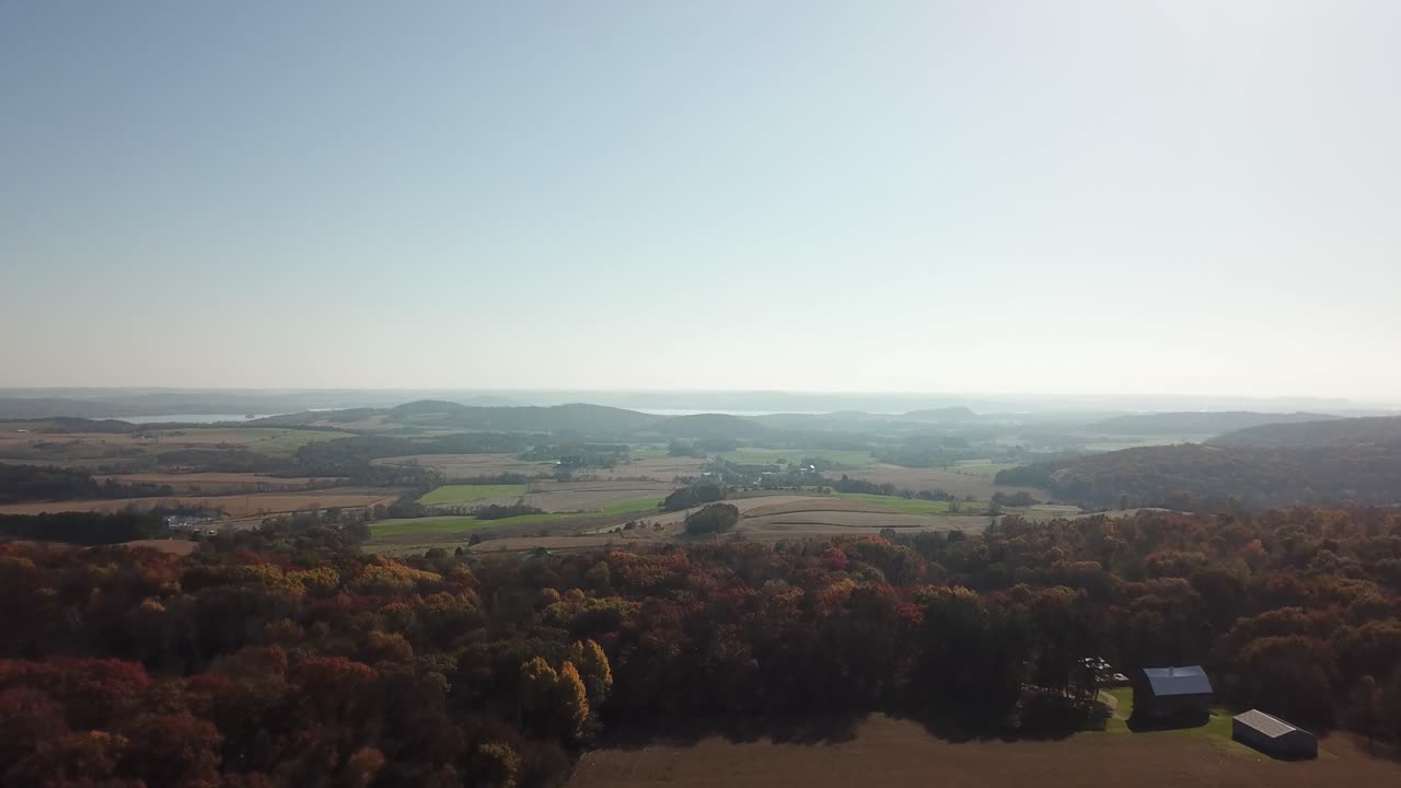 Expansive farmland bordered by vibrant autumn-colored forest, with a barn and hills in the background under a clear sky. Captures a serene rural Midwest landscape.