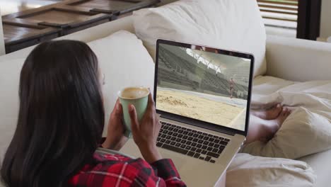 Composite-of-woman-sitting-at-home-holding-coffee-watching-athletics-long-jump-event-on-laptop