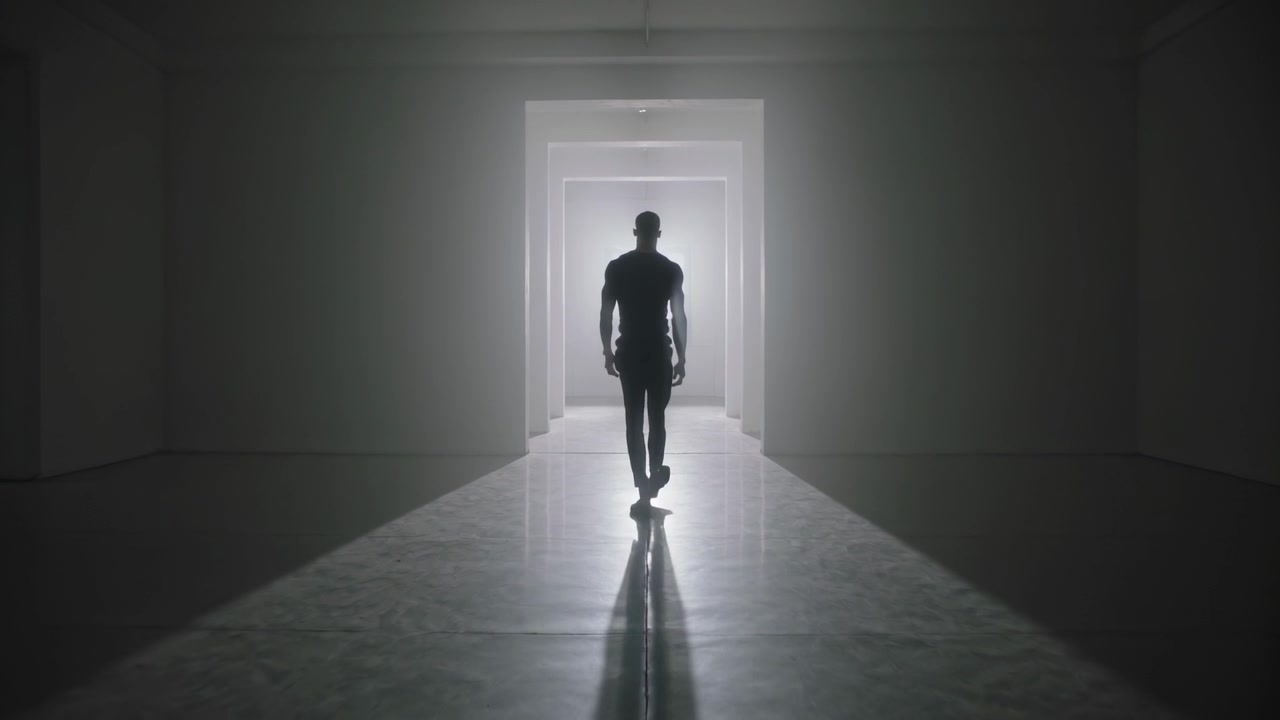 Sagoma maschile che cammina, emergendo da un portale luminoso, che avanza lungo il pavimento di un corridoio con abiti scuri.
