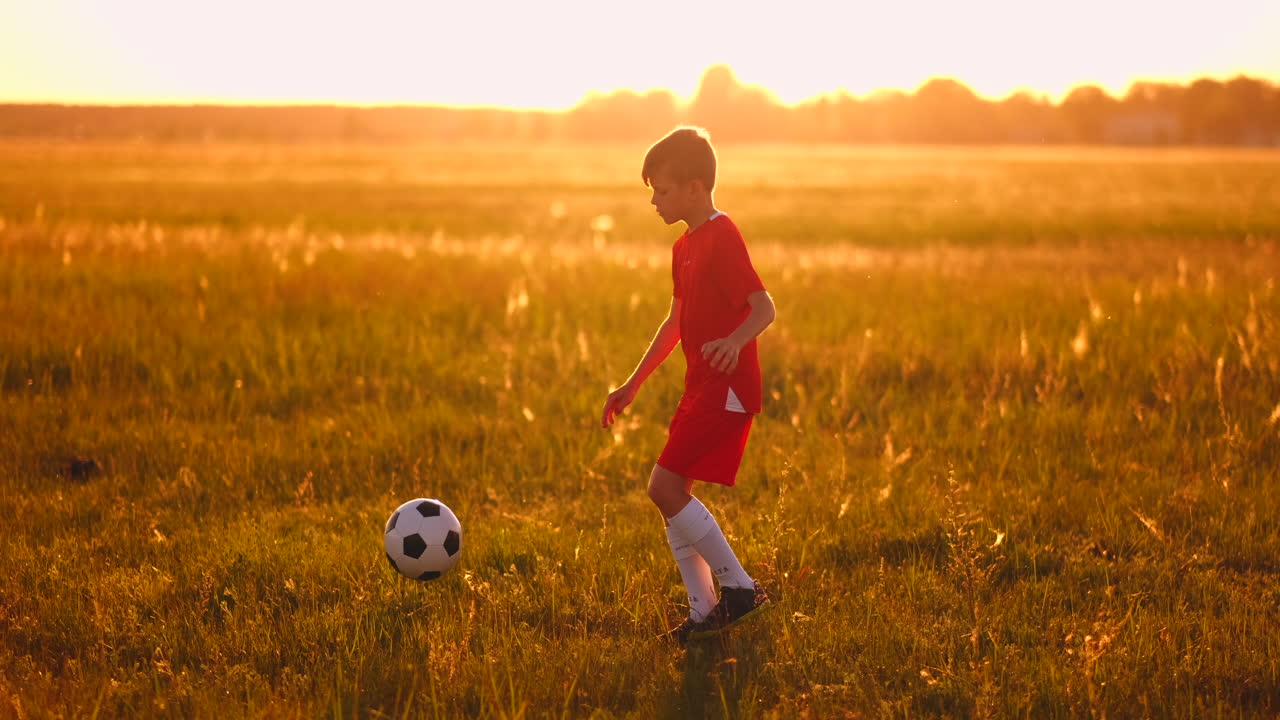 niño junior con una camiseta roja y zapatillas de deporte al atardecer haciendo malabarismos con un entrenamiento de pelota de fútbol y preparándose para convertirse en un jugador de fútbol. el camino hacia el sueño. trabajo duro