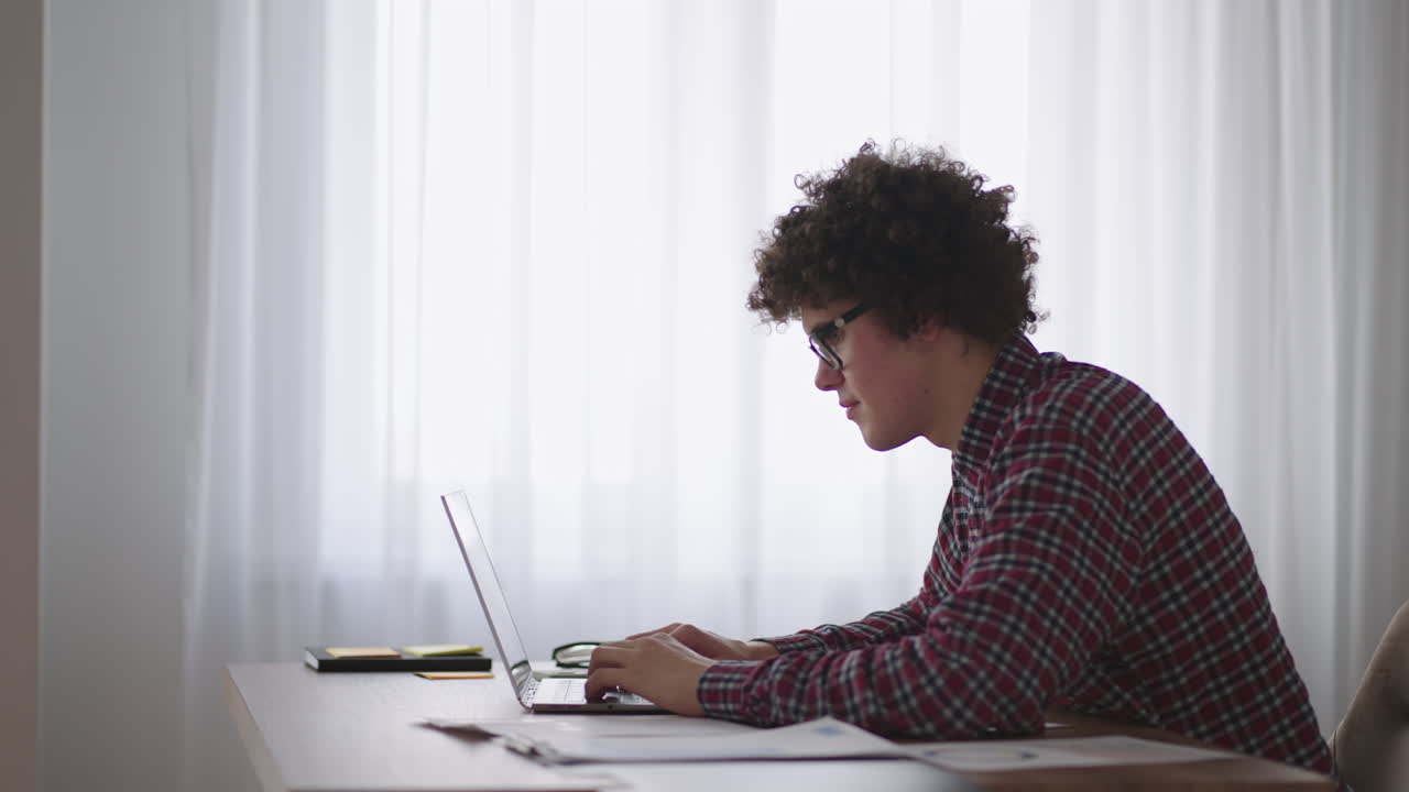 estudiante de cabello rizado atractivo joven con gafas está estudiando en casa usando una computadora portátil escribiendo en un cuaderno. estudiante universitario usando una computadora portátil viendo un seminario de aprendizaje en línea a distancia