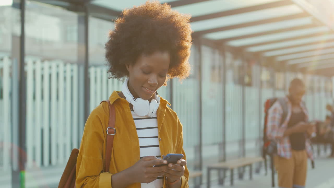 Young African American woman traveller with backpack texting message on smartphone and smiling cheerfully at train station