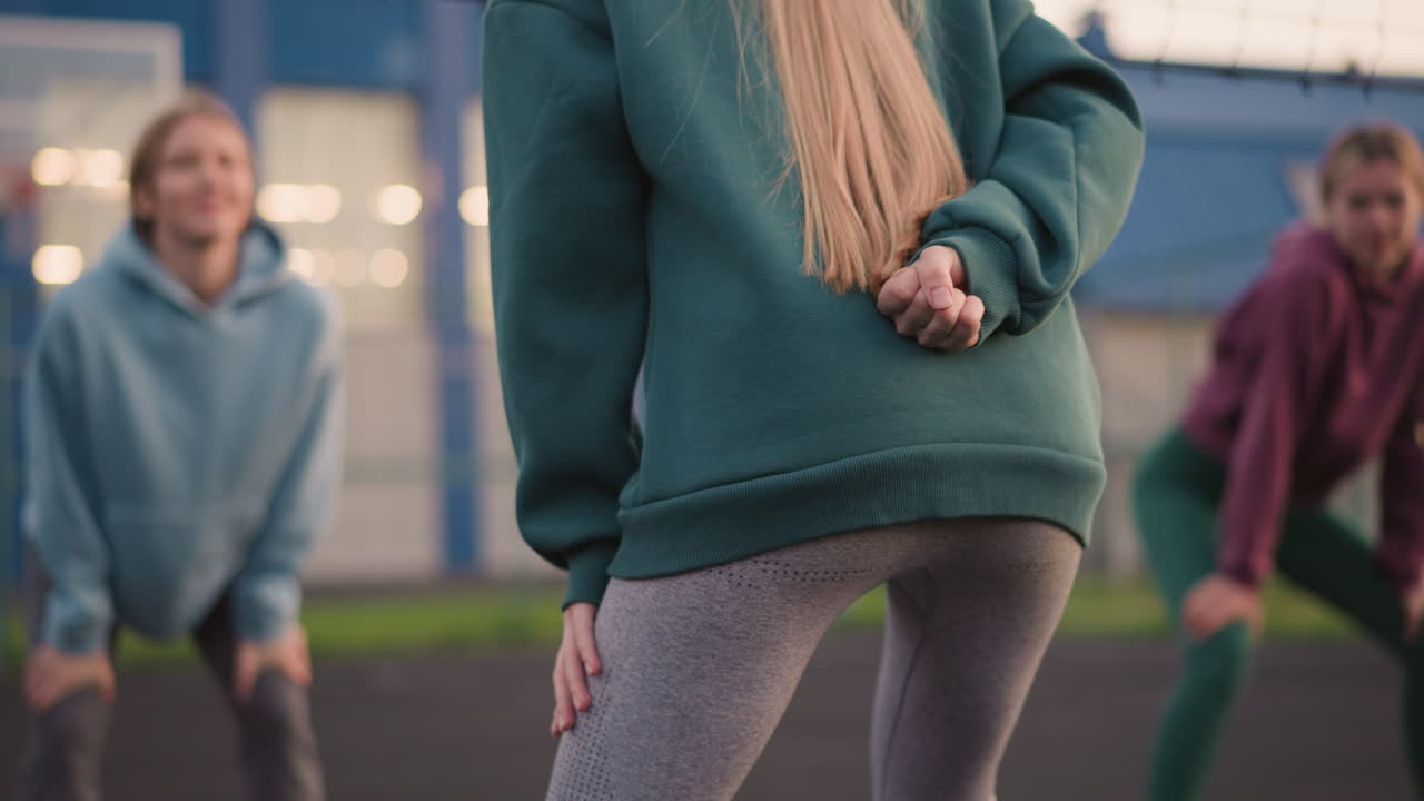 초록색 후디를 입고 몸짓을 하는 여성의 뒷면, 배경에 약간 구부러진 두 여성, 모두 야외 코트에서 신체 훈련에 종사하고 있습니다.