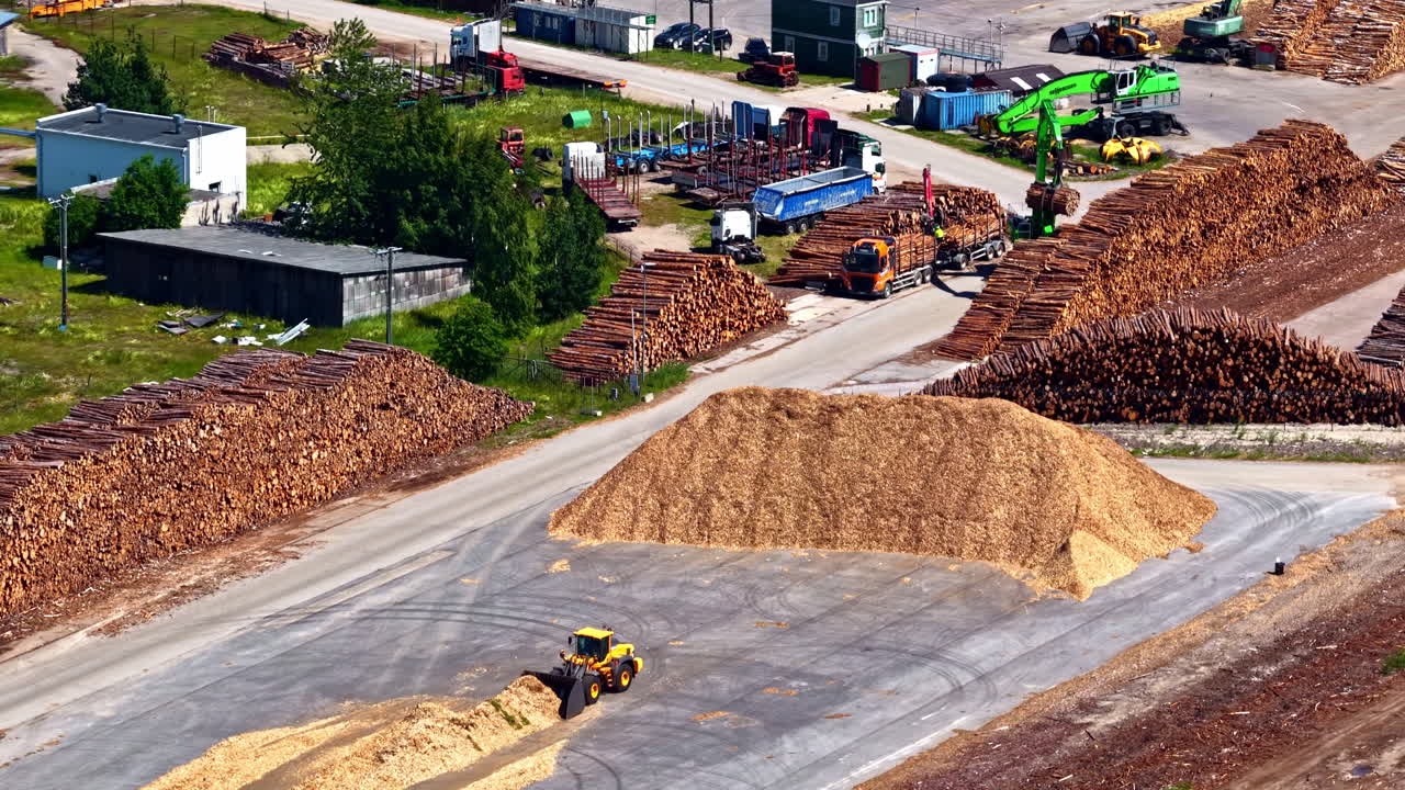 Aerial drone view over a vast logging factory in Latvia, showing stacks of raw timber, piles of wood chips, and heavy machinery like loaders and cranes at work in the wood processing industry