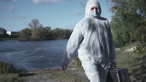 woman in face mask and protective suit holding case and walking out of river or water, industrial background