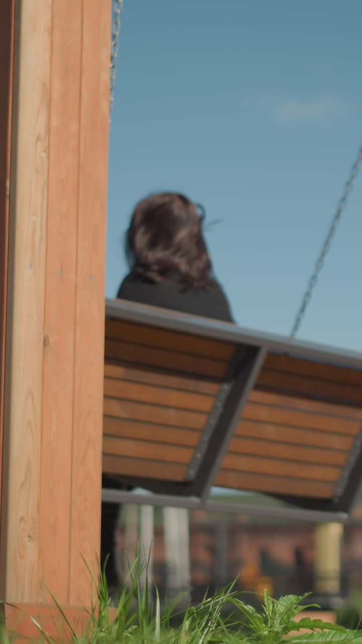 바람에 날아다니는 머리카락으로 흔들리는 스 의자에 앉은 여자, 배경에 있는 나무, 그리고 공립 공원에서 멀리서 흐릿한 사람