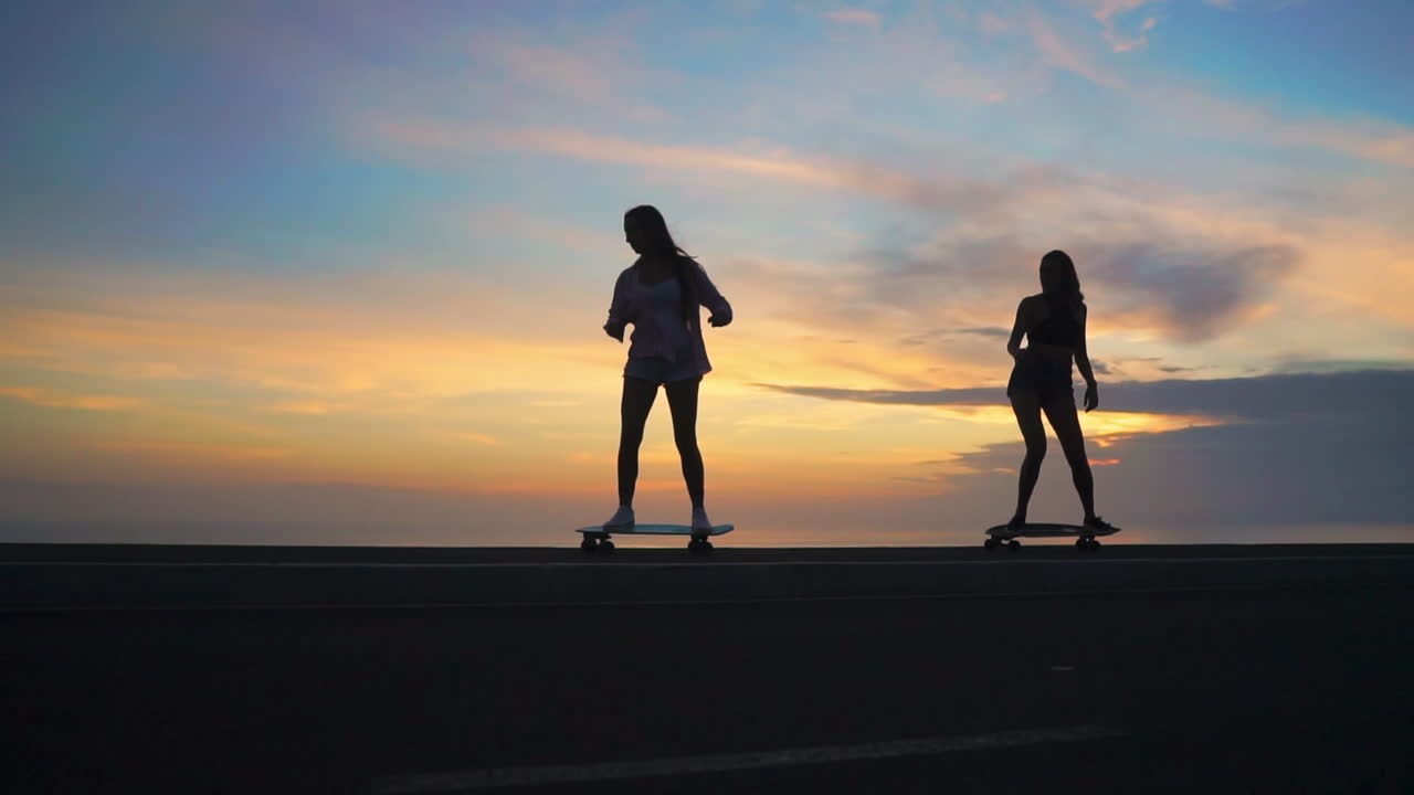 Two friends enjoy slow-motion skateboarding along a road during sunset, framed by mountains and a scenic sky. Their attire features shorts