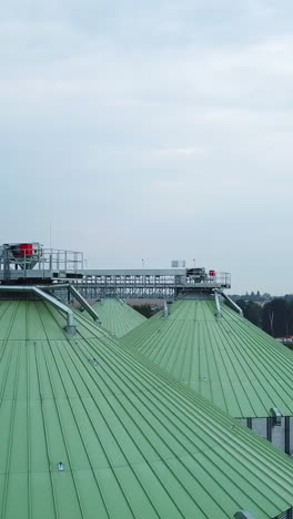 rising along a huge silver metal granary tank standing at the territory of modern silo plant. look at the slope roofs of elevators at the backdrop of farmlands. vertical video